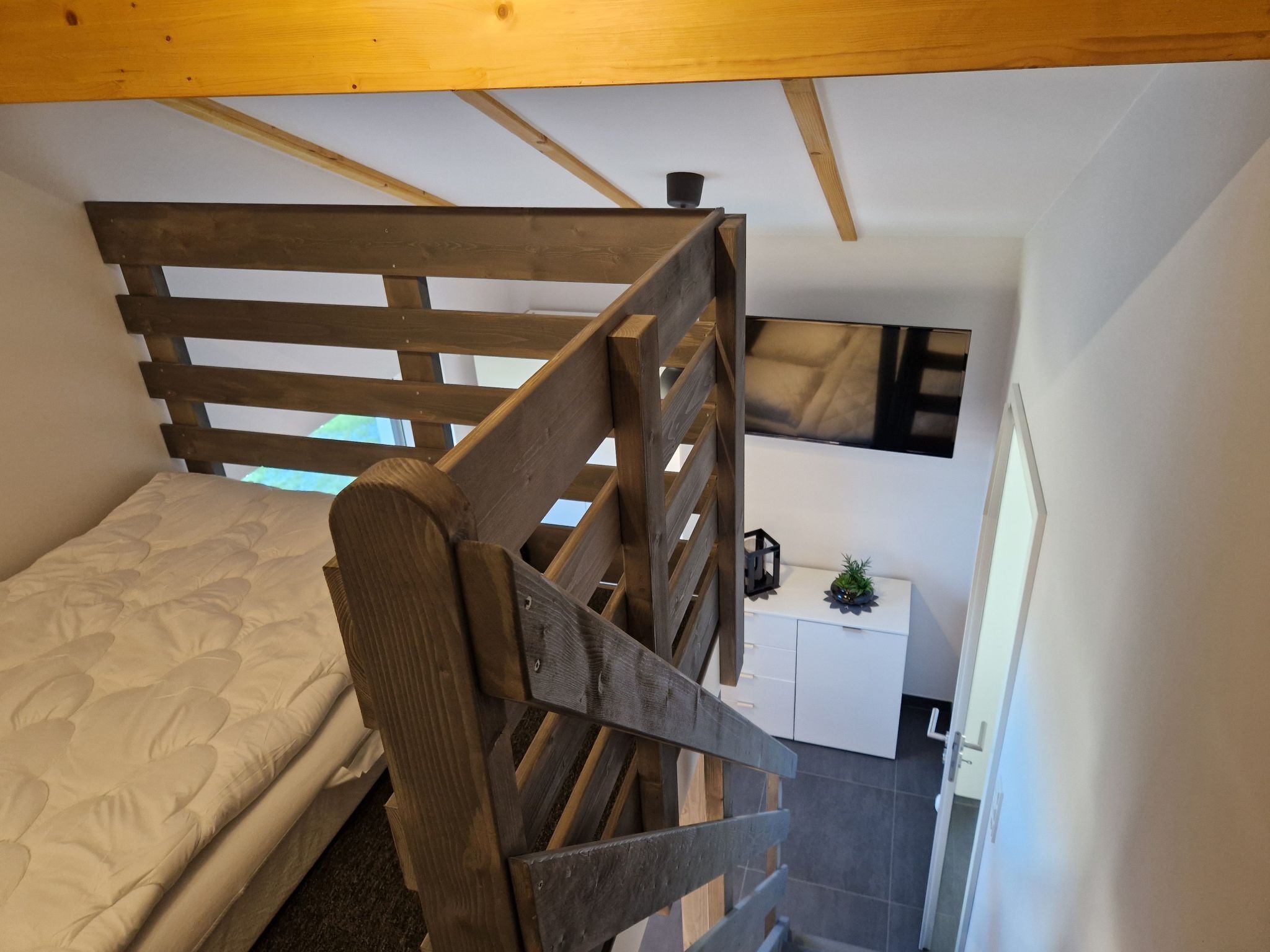 A bedroom with a wooden bunk bed, a white bedside table with a small plant, and a window with a view of the sky.