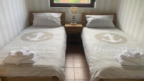 Zwei Einzelbetten mit passenden Kopfteilen und Fußteilen, jeweils mit einem zusammengerollten Handtuch und einem Kissen. Die Betten haben ein Design im Seestil mit Anker-Motiven. Darüber befindet sich ein Fenster, und dazwischen ein Nachttisch mit einer Lampe.
