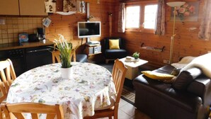 Een gezellig kamer in blokhutstijl met houten inrichting, een eettafel met bloemenonderzetter, een bank, een televisie en een raam met gordijnen.