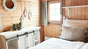 Un dormitorio acogedor con pared de madera, cama con ropa de cama blanca, una ventana pequeña y un tocador con espejo y adornos.