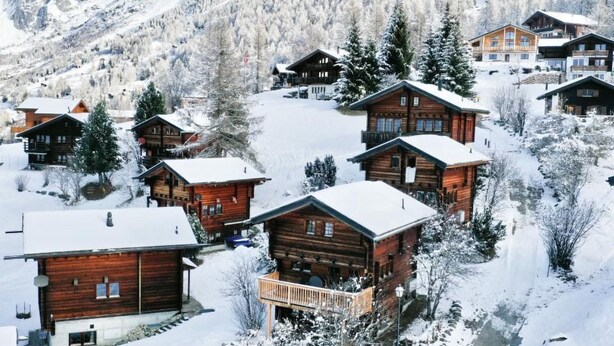 Un grupo de cabañas de madera con tejados nevados, rodeadas de árboles cubiertos de nieve y montañas al fondo.