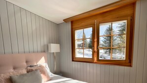 A bedroom with a bed, a nightstand, a lamp, and a window with a view of trees and snow. - Wohnung Vieux Valais by Interhome (Bellwald)
