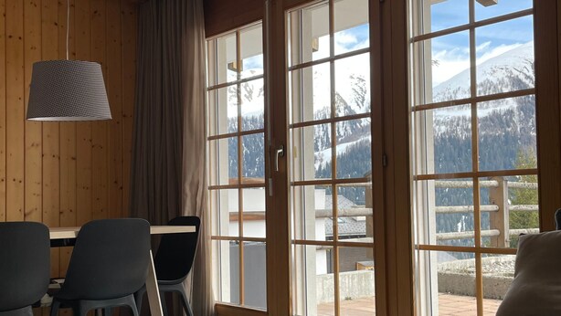 A room with a wooden paneled wall, a dining table with chairs, a sofa, and large windows with a mountain view. - Wohnung Vieux Valais by Interhome (Bellwald)