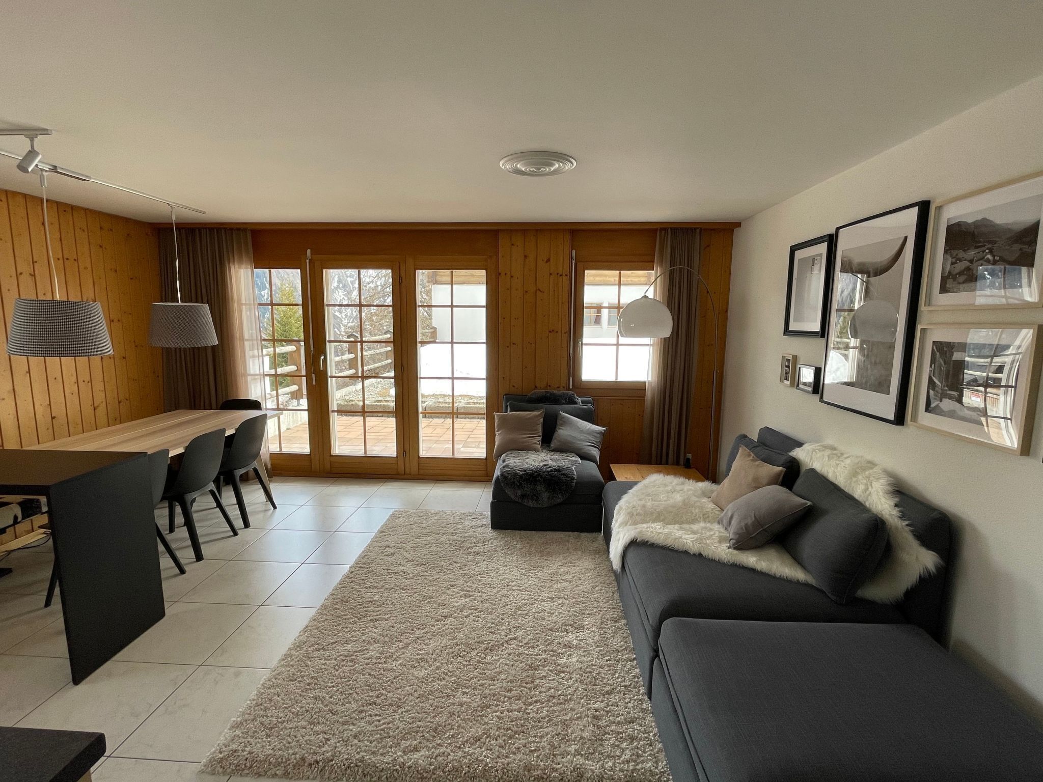 Uma sala de estar moderna com piso de madeira, um sofá cinza, uma mesa de jantar com cadeiras e uma janela grande com vista para o exterior.
