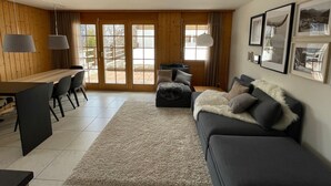 A modern living room with a wooden floor, a grey sofa, a dining table with chairs, and a large window with a view of the outdoors. - Wohnung Vieux Valais by Interhome (Bellwald)