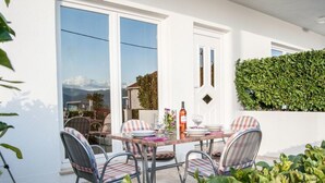 Un patio al aire libre con una mesa dispuesta para comer, rodeado de sillas, una pared blanca y un seto verde.