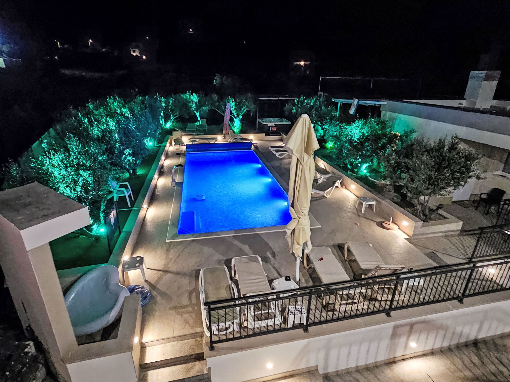 A rooftop pool with a blue pool, surrounded by lounge chairs and umbrellas, and illuminated by green lights.