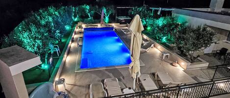 A rooftop pool with a blue pool, surrounded by lounge chairs and umbrellas, and illuminated by green lights.