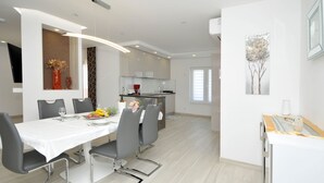 A modern kitchen with white cabinets, a dining area with a white table and grey chairs, and a small dining table with a fruit bowl and flowers. - Villa Rimac in Kastel Stafilic mit Garten, Grill by Interhome (Kaštel Novi)