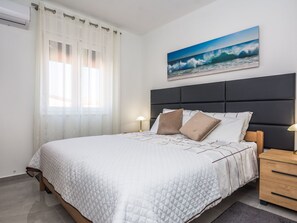 Ein ordentlich eingerichtetes Schlafzimmer mit einem Bett, einem Kopfteil, einem Nachttisch, einer Lampe und einem Bild mit einer Strandansicht.