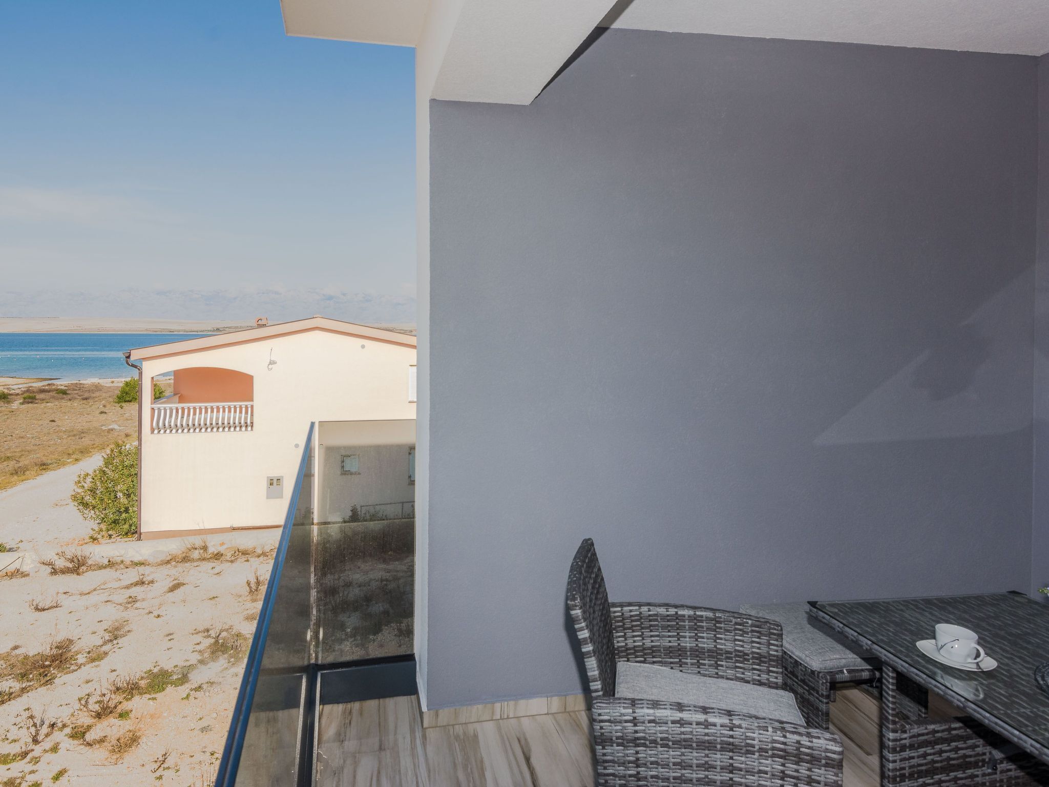 Ein Balkon mit Blick auf Strand und Meer, ein Esstisch mit Stühlen und ein kleines Gebäude im Hintergrund.