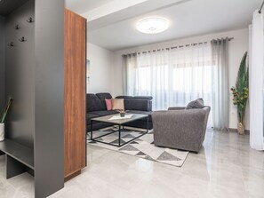 Ein modernes Wohnzimmer mit einem dunklen Sofa, einem grauen Sessel und einem Couchtisch. Ein großes Fenster mit durchsichtigen Vorhängen bietet Blick auf die Stadt.