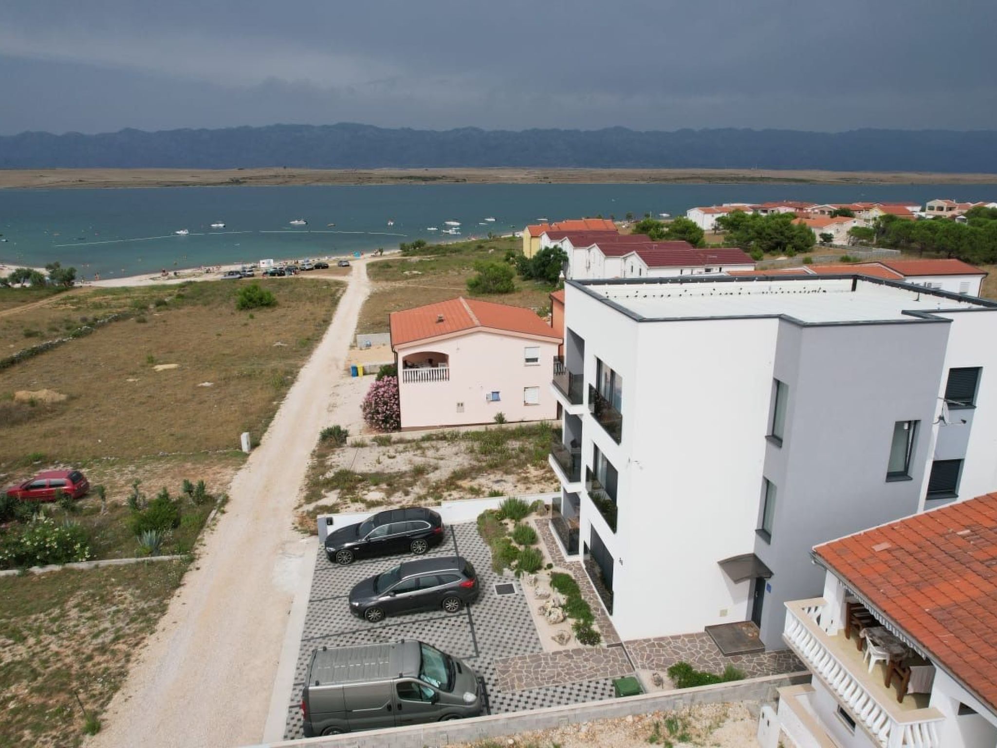 A coastal building with a parking area, a beach in the background, and mountains in the distance.