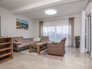 Ein modernes Wohnzimmer mit Sofa, Sesseln und Couchtisch. An der Wand hängt ein Gemälde, eine Pflanze steht dort und es gibt ein Fenster mit Vorhängen.