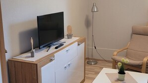 Ein modernes Wohnzimmer mit einem Flachbildfernseher auf einem Holzschrank, einem weißen Couchtisch mit einer Pflanze, einem beigen Sofa und einer Stehlampe.