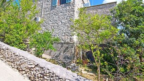 Un bâtiment en pierre, doté d’une petite fenêtre et de volets, entouré de verdure et d’un mur de pierre.