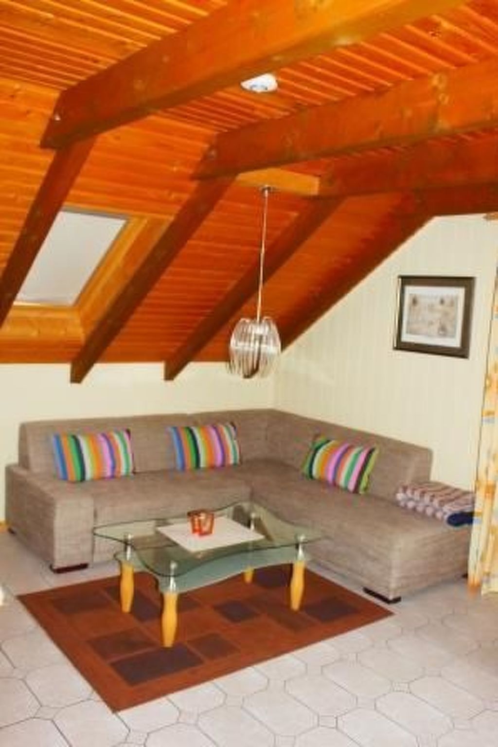 A modern living room with a glass coffee table, a gray sectional sofa with colorful cushions, wooden beams on the ceiling, and a skylight.