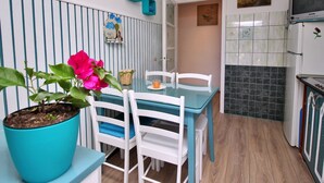 Een keuken met een betegelde achterwand, een eethoek met een blauwe tafel en witte stoelen, en een potplant met roze bloemen.