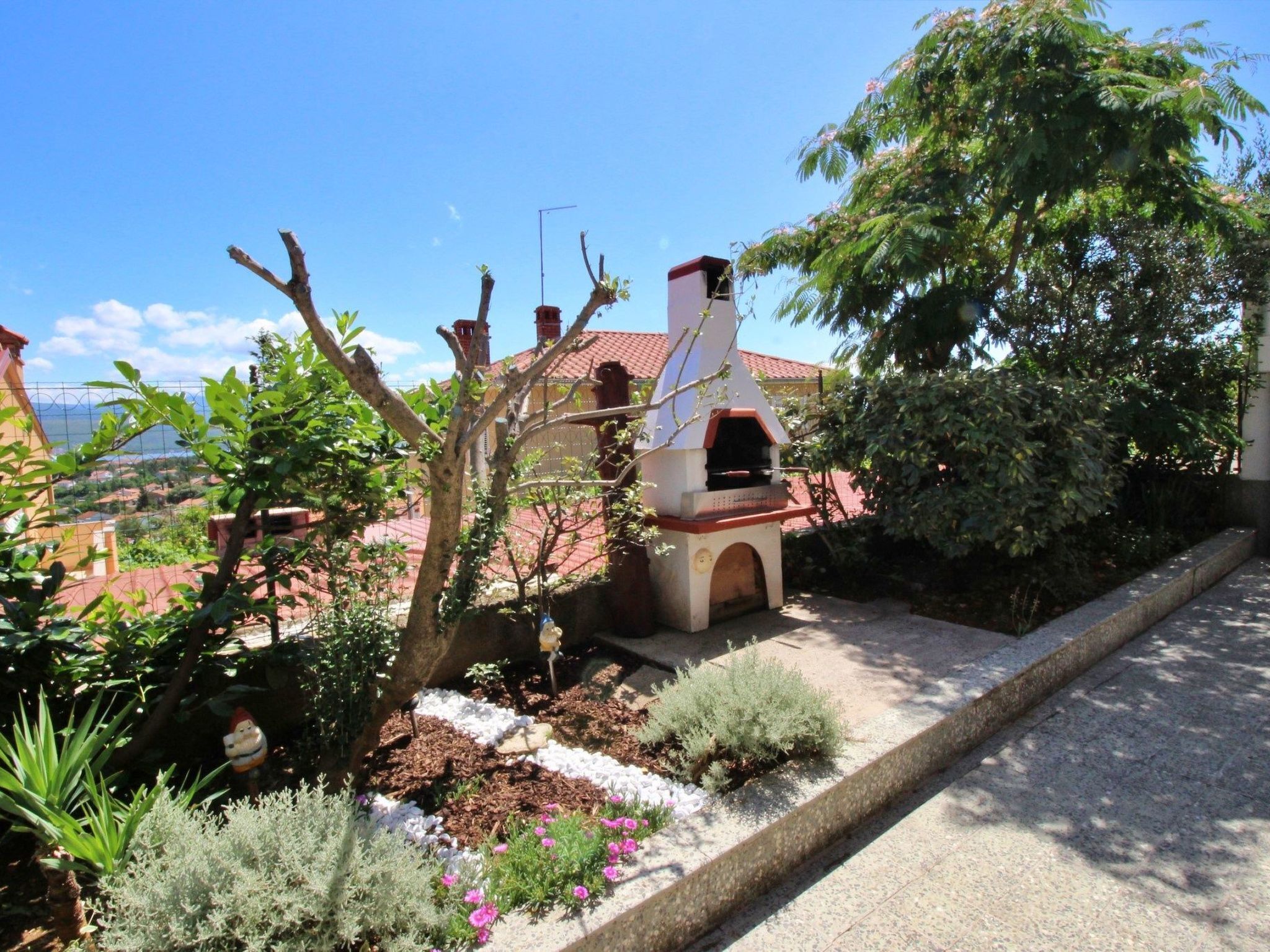 A garden with a barbecue area, a decorative gnome, and a view of a red-roofed building in the background.