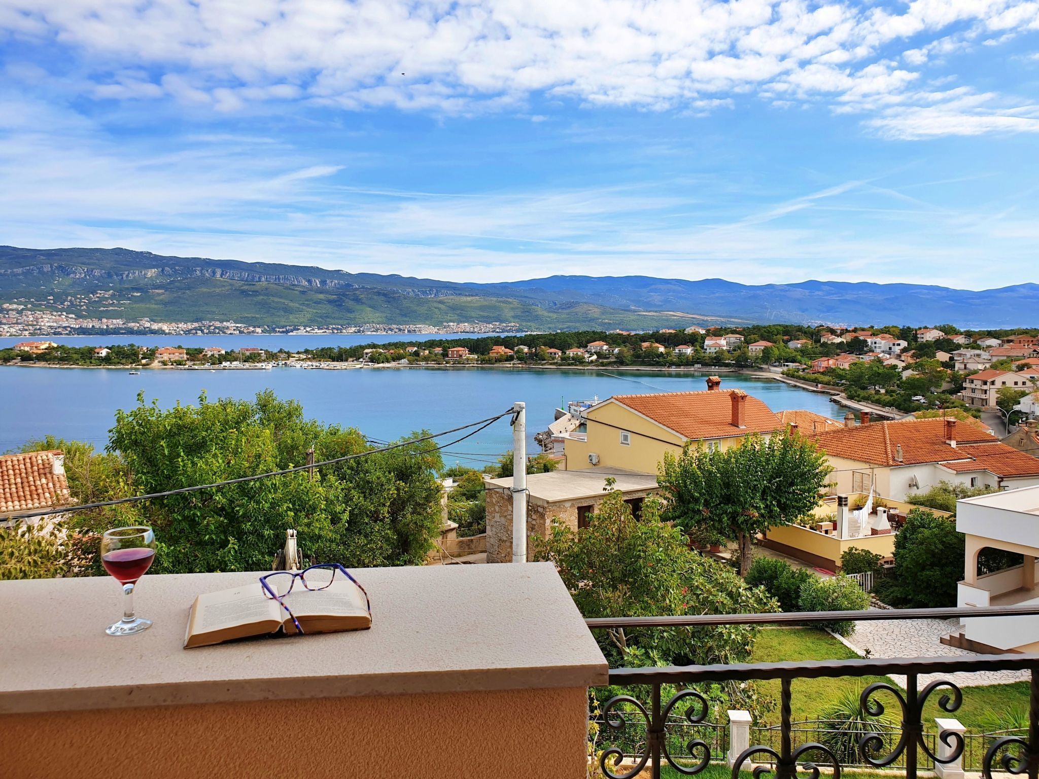 Un balcon avec un livre et un verre de vin rouge, donnant sur un lac, avec des maisons et des montagnes en arrière-plan.