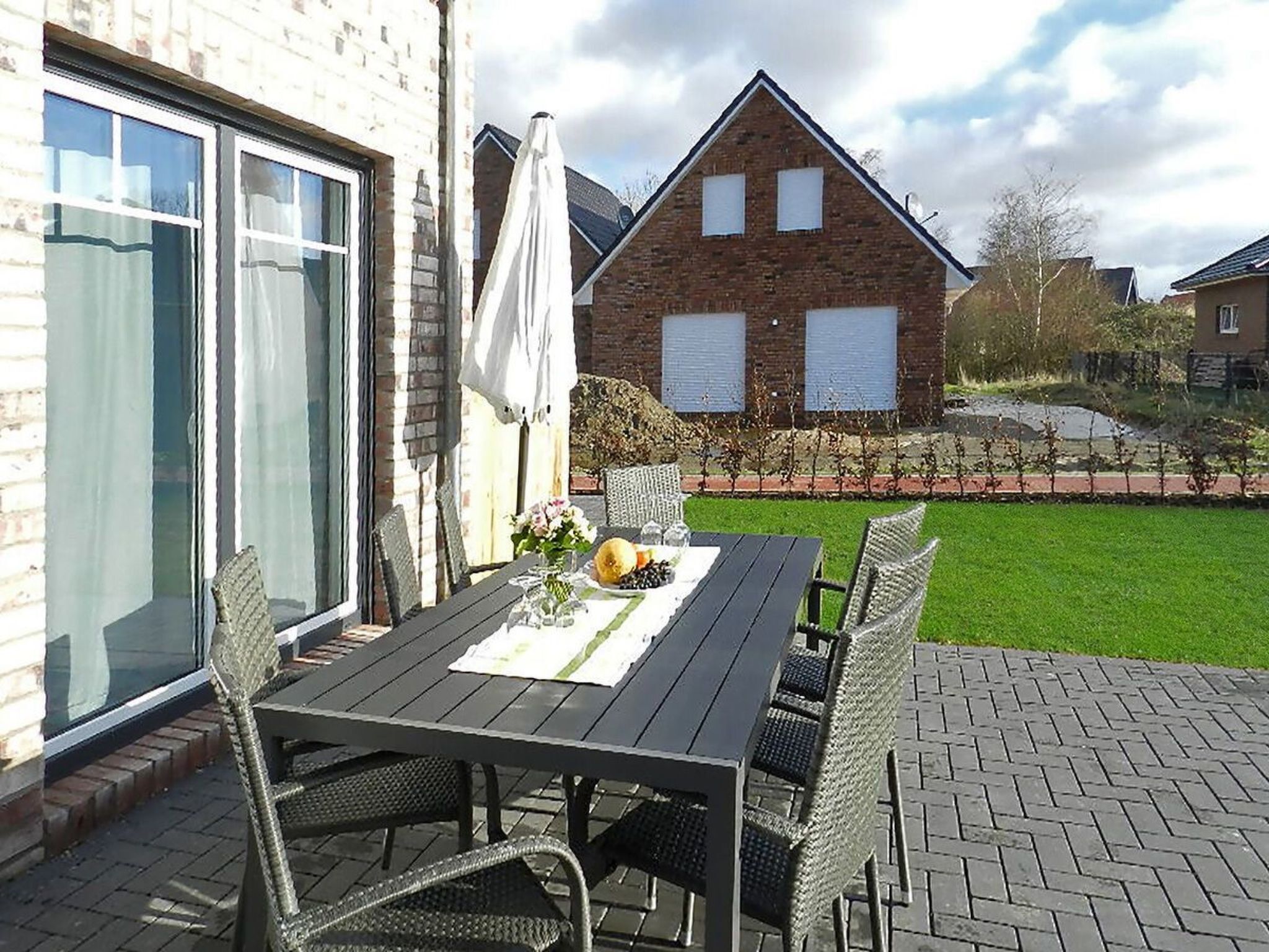 Eine Terrasse mit Tisch und Stühlen, im Hintergrund ein Backsteinhaus und eine Rasenfläche.