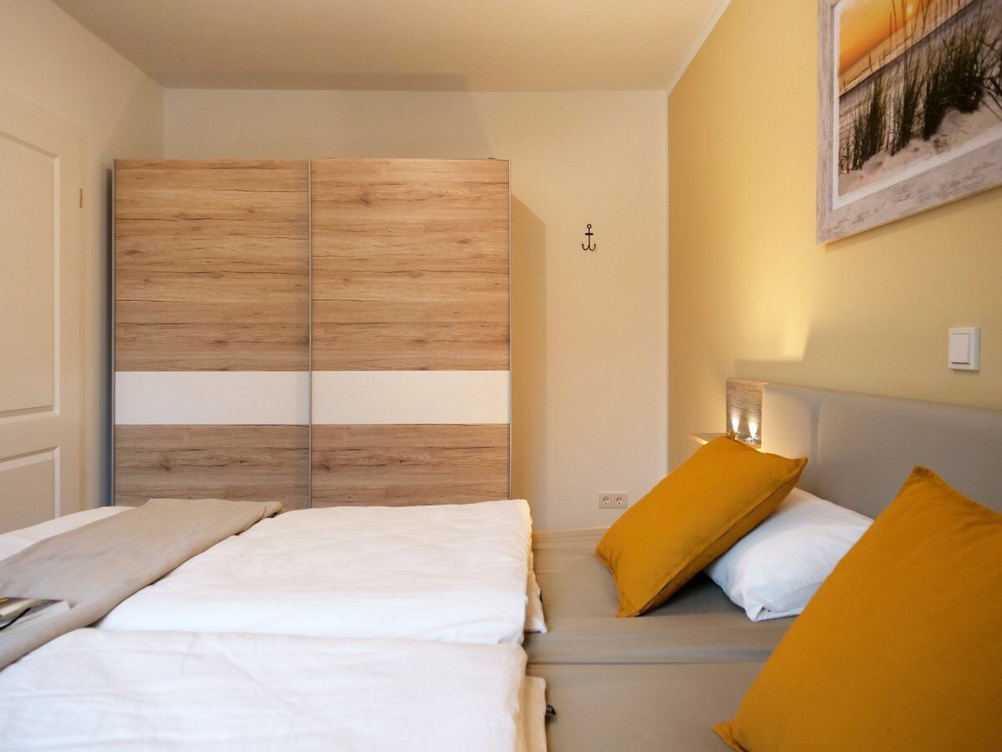 A bedroom with a wooden wardrobe, a bed with white and mustard pillows, and a framed picture on the wall.