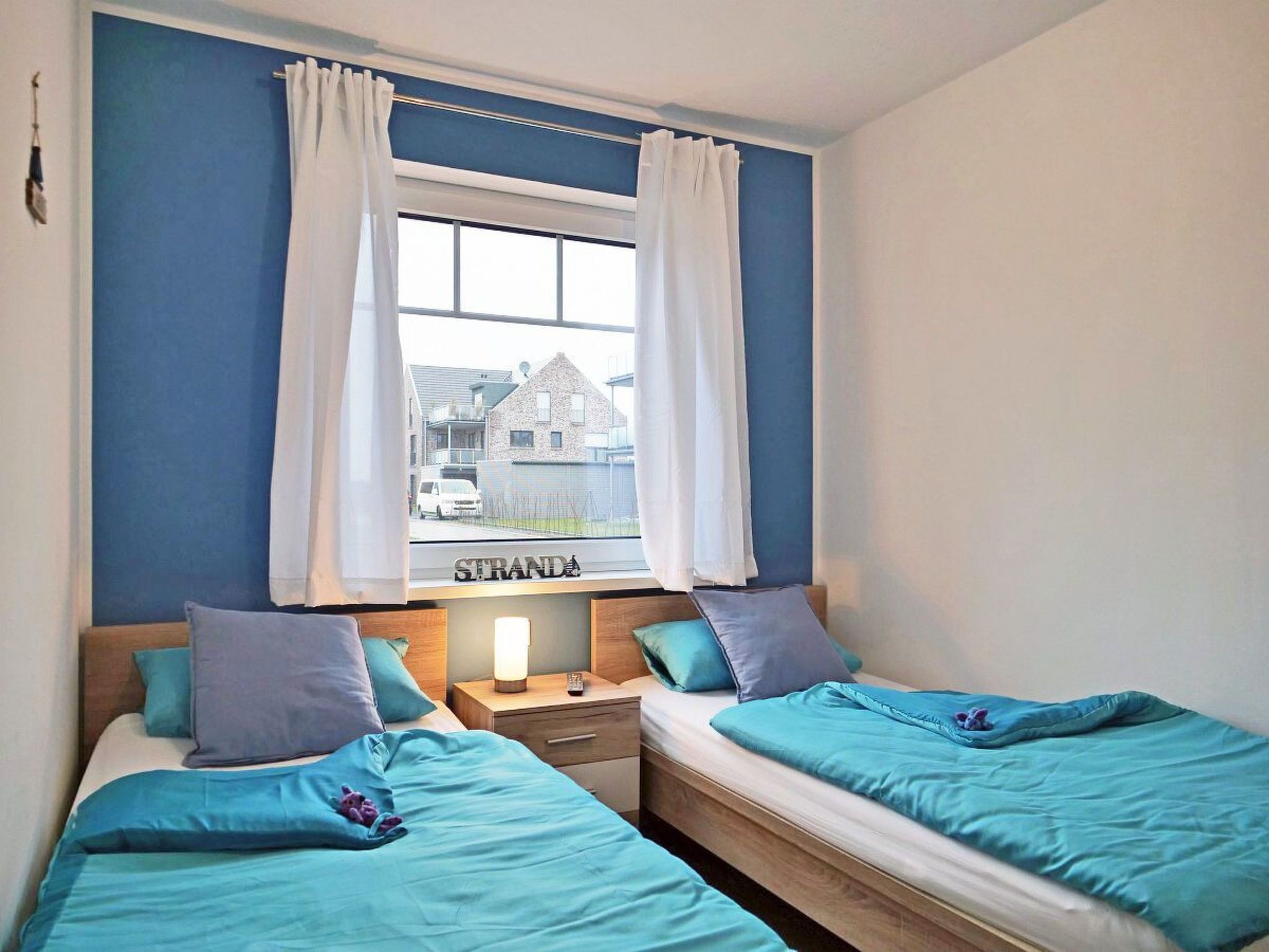 A bedroom with two beds, a window with curtains, a bedside table with a lamp, and a view of a street outside.