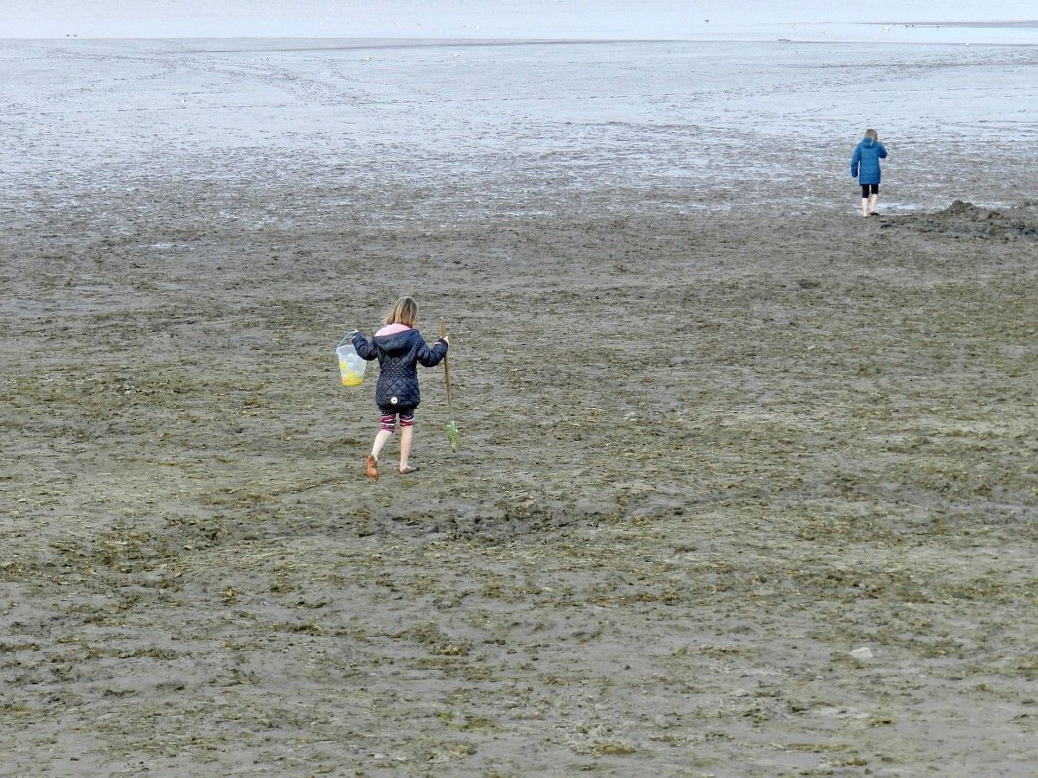 A beach scene with two individuals walking away from the camera, one carrying a bucket.