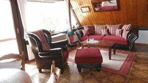 Un salon chaleureux avec un canapé en cuir, une table basse en bois et un pouf rouge. On y trouve également une chaise en bois et une petite table sur laquelle sont posés un vase et des verres. Les murs de la pièce sont en bois et elle est dotée d’une grande fenêtre.