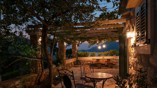 Ein Patio im Freien mit Steinböden, einem runden Tisch mit Stühlen, einer Pergola mit Lichterketten und Blick auf die Berge im Hintergrund.