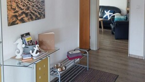 Een moderne woonkamer met een glazen bureau, een schilderij van een strand, een houten deur en een zicht op een andere kamer met een bank en een schilderij.