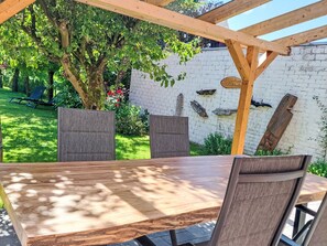 Ein Tisch aus Holz mit Stühlen, eine Pergola, eine weiße Backsteinmauer und ein Baum mit grünen Blättern.