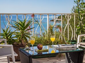 Uma mesa de vidro com bebidas e um livro, cercada por plantas, com vista para o mar.