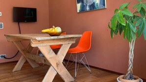 Uma mesa de jantar de madeira com uma tigela de frutas, cadeiras laranjas, uma planta em vaso e um quadro na parede.