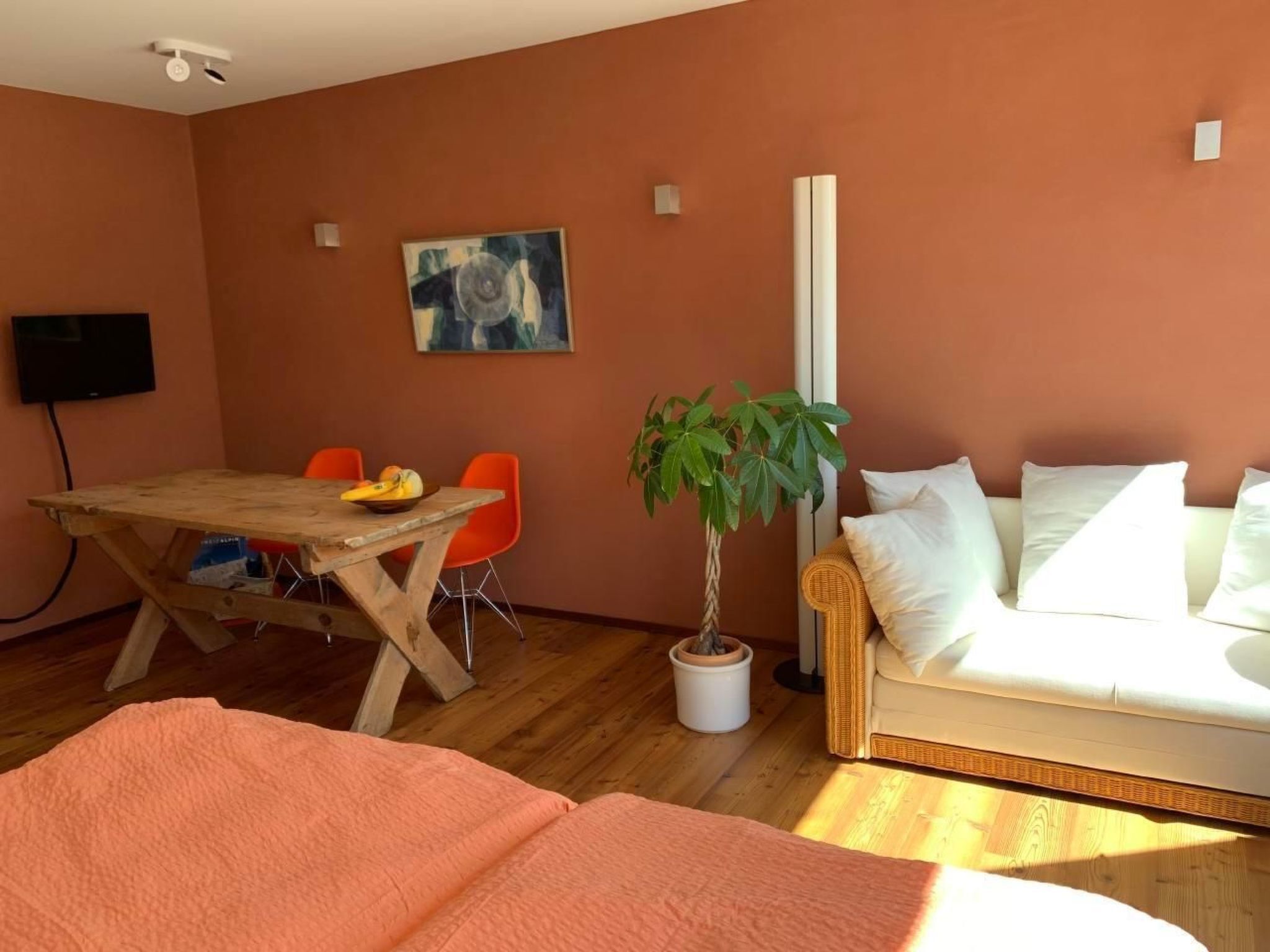 A room with a wooden dining table, orange chairs, a sofa, a potted plant, and a painting on the wall.
