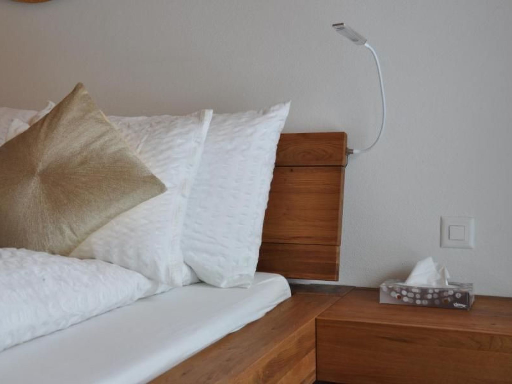 Un lit bien fait, avec du linge de lit blanc et un seul oreiller marron, une table de chevet en bois avec une boîte de mouchoirs, et une prise électrique fixée au mur.