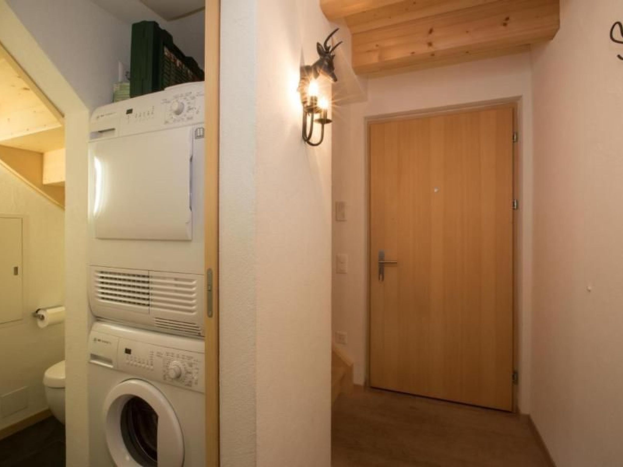 A laundry room with stacked appliances, a wooden door, and a wall-mounted light fixture.