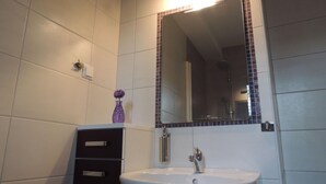 Een badkamer met een wit wastafelmeubel, een spiegel met een betegelde lijst, en een kleine paarse vaas op het aanrecht.