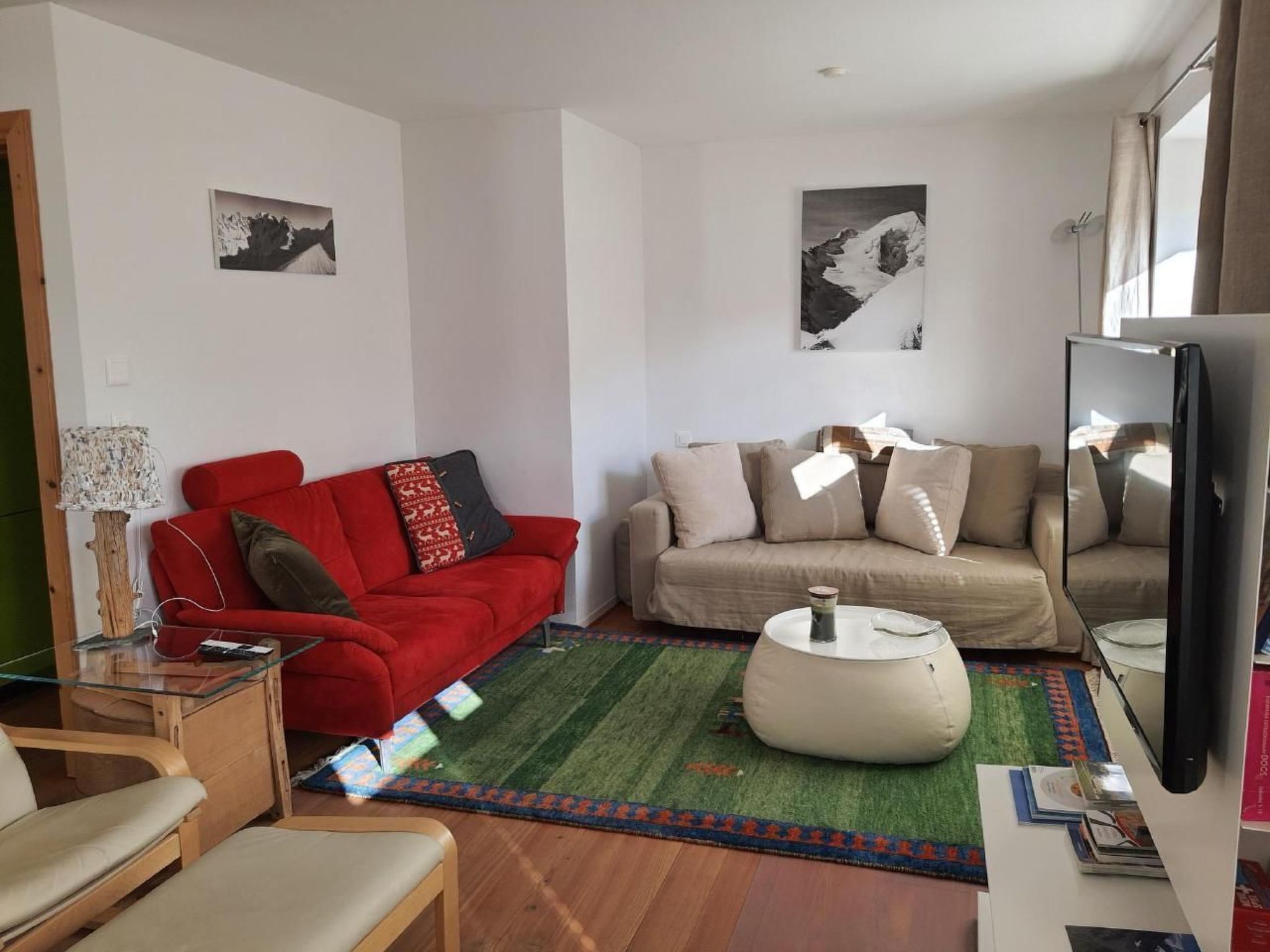 En stue med en rød sofa, en beige sofa og et sofabord i glas. Der hænger to billeder på væggene, og et fjernsyn er placeret til højre.