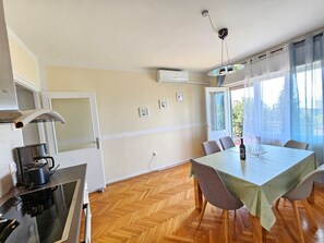 Moderni keittiö, jossa on liesi, uuni ja kahvinkeitin. Keittiössä on ruokapöytä tuoleineen, ja suurista ikkunoista avautuu näkymä ulos.