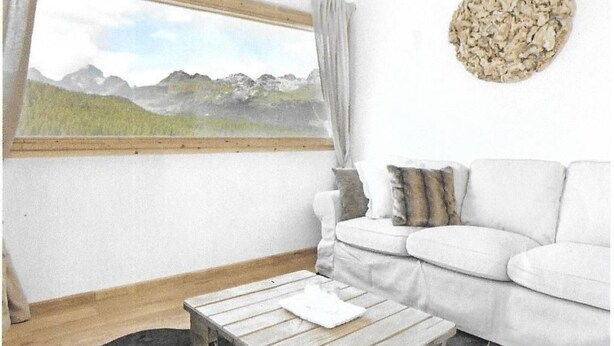 Una habitación con un sofá blanco, una mesa de centro de madera, una ventana con vistas a montañas y una decoración en la pared.