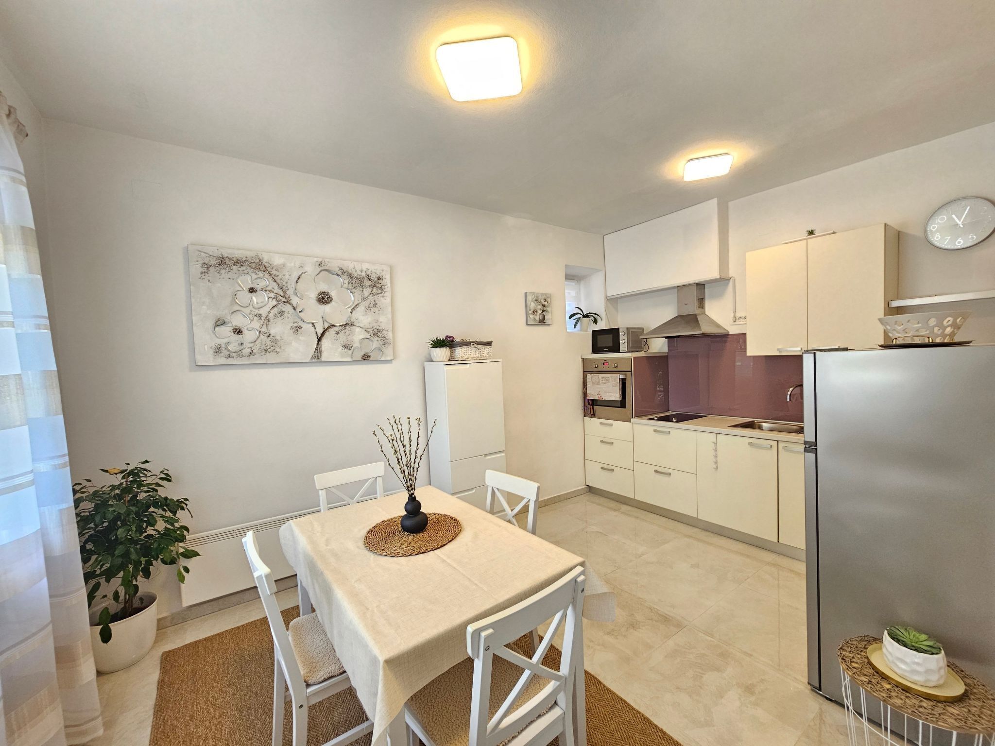 A modern kitchen with white cabinets, a stainless steel refrigerator, and a dining area with a table and chairs.