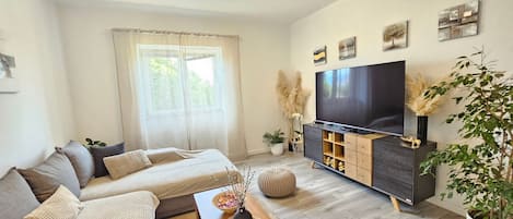 A modern living room with a sofa, coffee table, TV stand, and a large window with sheer curtains.