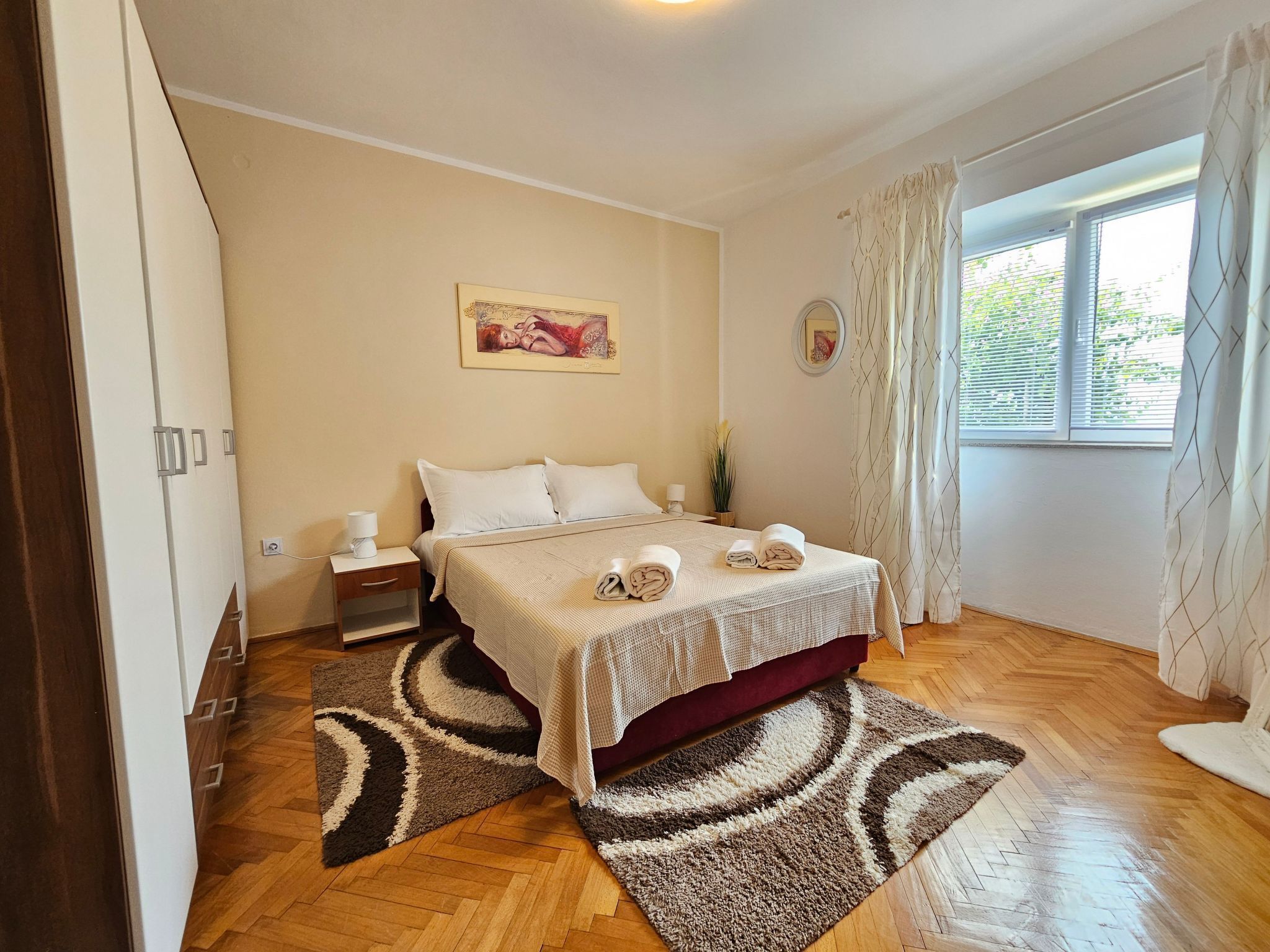 A bedroom with a bed, a nightstand, a lamp, a painting on the wall, and a window with curtains.
