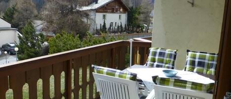 A balcony with white plastic chairs and a table, overlooking a mountainous landscape with a house in the background.