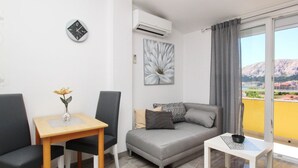 Ein modernes Wohnzimmer mit einem grauen Sofa, zwei schwarzen Sesseln, einem Holztisch mit Blumen und einem weißen Tisch mit einer Schüssel. An der Wand hängt ein Gemälde, über dem Sofa befindet sich eine Klimaanlage.