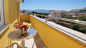 Ein Balkon mit Tisch und Stühlen, einer Obstschale und Blick auf eine Küstenstadt und Berge.