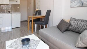 Ein modernes Wohnzimmer mit grauem Sofa, gemusterten Kissen, einem Couchtisch mit Schale, einem Essbereich mit Holztisch und Stühlen sowie einer Küche im Hintergrund.