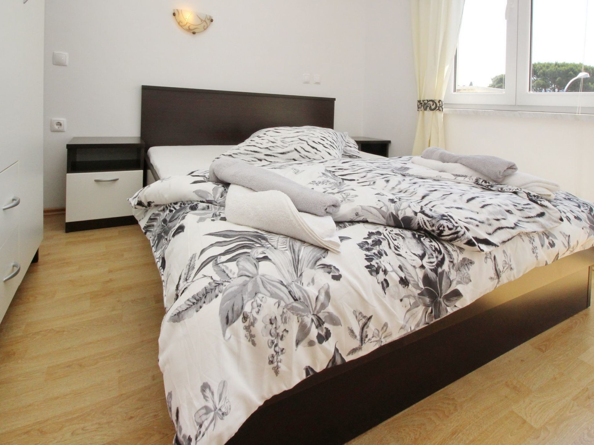 Ein ordentlich bezogenes Bett mit floralem Überwurf, ein Nachttisch mit Lampe und ein Fenster mit Vorhängen.
