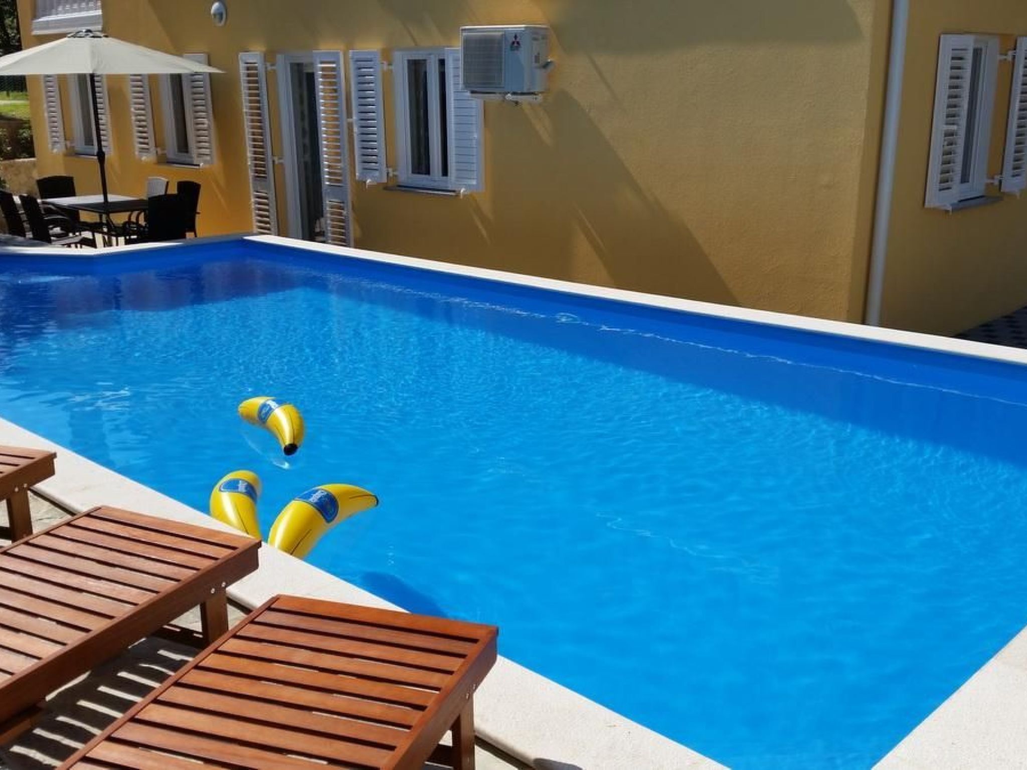 Uma piscina com espreguiçadeiras de madeira e uma mesa, rodeada por paredes amarelas com persianas brancas.